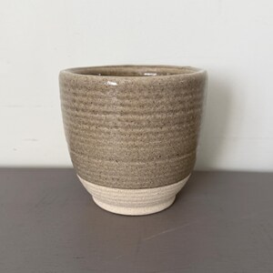 May include: A handcrafted ceramic cup. The upper half is light brown with a textured finish, the base is cream. The cup has a rounded shape and a minimalist design. Ideal for drinks or small items.