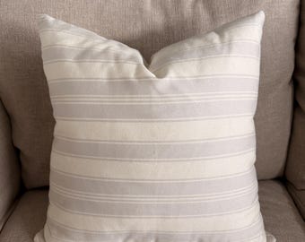 May include: A square cushion with horizontal stripes in light grey and off-white. The cushion is placed on a beige sofa, creating a neutral aesthetic. The cushion's fabric appears soft and textured, suitable for home decor.