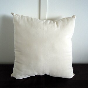 May include: A square, off-white pillow with a soft, textured appearance. The pillow has a simple design and is likely made of a natural fabric. The pillow is placed on a dark surface, and the background is a white wall.