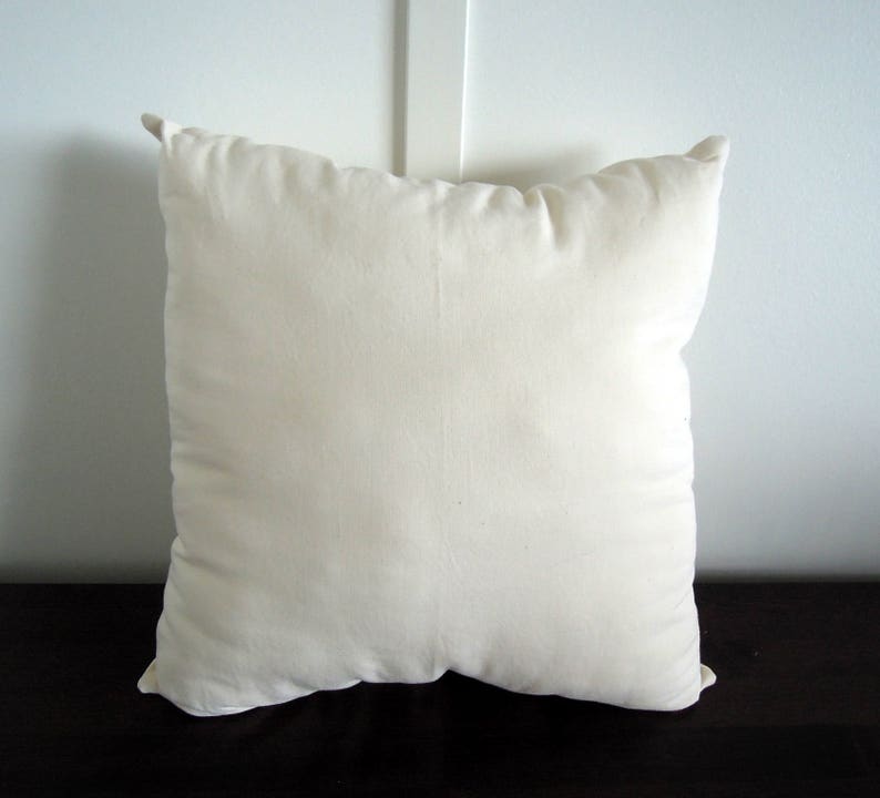 May include: A square, off-white pillow with a soft, textured appearance. The pillow has a simple design and is likely made of a natural fabric. The pillow is placed on a dark surface, and the background is a white wall.