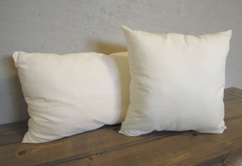May include: Two square, off-white pillows rest on a dark wooden surface. The pillows are soft and plush, with a slightly textured appearance. The background is a light gray wall, providing a neutral backdrop for the pillows.