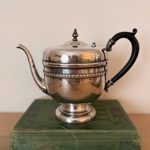 May include: A silver teapot with a black handle and a decorative lid. The teapot is on a vintage green book. The teapot has a shiny, reflective surface and a spout for pouring.