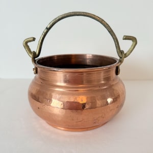 May include: A small copper cauldron with a hammered finish. It features a rounded body and a brass handle. The handle arches over the top. The cauldron is likely for decorative use or small-scale cooking.