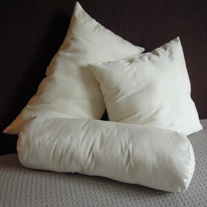 May include: Four ivory-colored pillows of varying shapes and sizes. There are two square pillows, one triangular pillow, and a cylindrical bolster pillow. The pillows are arranged on a gray textured surface, with a dark brown background.