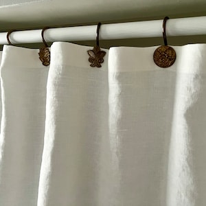 May include: White linen curtains suspended from a rod with decorative bronze-coloured hooks. The hooks display diverse designs, including a fleur-de-lis and floral patterns. The curtains are softly draped, forming gentle folds.