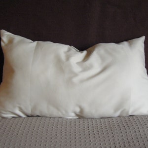 May include: A rectangular, off-white pillow with a soft, slightly wrinkled texture. The pillow is placed on a textured, light brown surface, with a dark brown background.