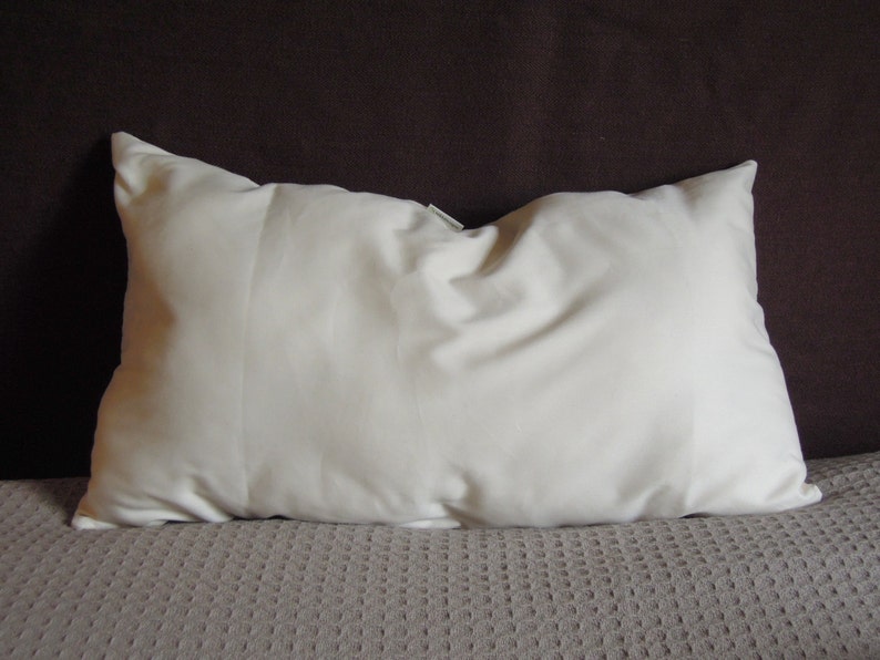 May include: A rectangular, off-white pillow with a soft, slightly wrinkled texture. The pillow is placed on a textured, light brown surface, with a dark brown background.