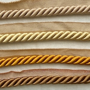 May include: Close-up of four decorative trims in varying shades of beige, gold, and brown. Each trim features a twisted rope design above a band of woven fabric. The trims are arranged horizontally against a textured white background.