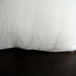 May include: Close-up of a cream-colored fabric, possibly linen or cotton, with a slightly textured surface. The fabric is curved at the bottom, suggesting a pillow or cushion. The background is a dark, wooden surface.