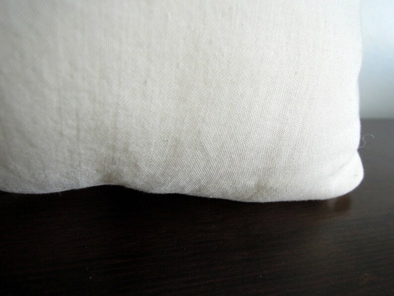 May include: Close-up of a cream-colored fabric, possibly linen or cotton, with a slightly textured surface. The fabric is curved at the bottom, suggesting a pillow or cushion. The background is a dark, wooden surface.