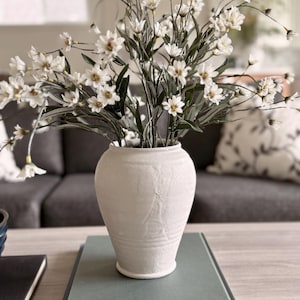 May include: A white ceramic vase filled with white flowers and green stems sits on top of a stack of books. The vase has a textured surface and a classic shape. The books are different shades of blue and green.