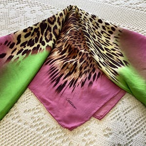 May include: A square silk scarf with a leopard print design. The scarf features a gradient of colors, including pink, green, and brown. The scarf is folded and displayed on a white lace surface.