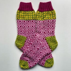 Op de afbeelding: Een paar kleurrijke gebreide sokken met een geometrisch patroon. De sokken hebben een magenta boord, een geel en groen geruit gedeelte, een magenta en wit ruitpatroon en groene hielen en tenen. De sokken zijn handgemaakt.