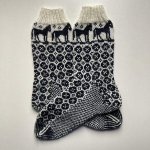 Op de afbeelding: Een paar gebreide sokken met een witte boord en een zwart-wit patroon. De sokken hebben een herhalend paardenontwerp aan de boord en een geometrisch patroon op de body. De sokken zijn gemaakt van een zacht, warm materiaal.