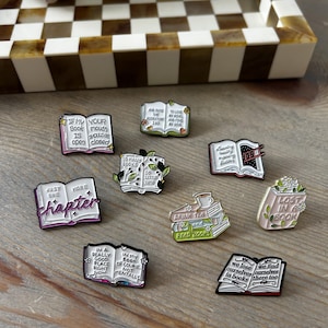 May include: A collection of nine enamel pins shaped like open books, each featuring unique illustrations and text. The pins display various literary quotes and designs, including stacks of books, tea cups, and floral accents in a variety of colours.