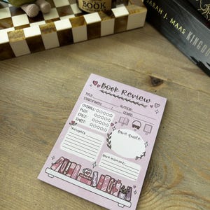 May include: A rectangular light purple notepad for book reviews featuring printed fields for title, author, start and finish dates, and genre. It includes star rating sections for overall, plot, spice, and sweet, plus spaces for thoughts, a favourite quote, and a favourite moment, with a bottom illustration of a bookshelf.