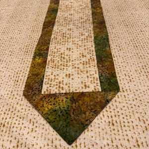 May include: A green, brown, and yellow patterned table runner with a triangular shape. The runner is laid on a white tablecloth with a pattern of small brown dots.