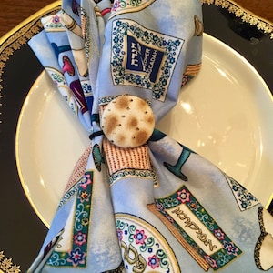 May include: A blue and white fabric napkin with a Passover theme. The napkin is folded and tied with a round, beige matzah cracker. The napkin features Hebrew text and images of traditional Passover symbols.