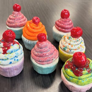 Può includere: Una collezione di sculture di cupcake in ceramica colorate. Ogni cupcake presenta un design di glassa a spirale in vari colori, sormontato da una ciliegia rossa brillante. I cupcake sono esposti su una superficie scura.