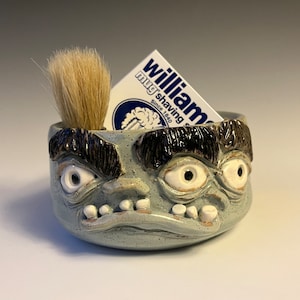 Puede incluir: Un tazón de afeitar de cerámica con una cara de tres ojos, una brocha de afeitar tupida y una tarjeta de jabón de afeitar William.
