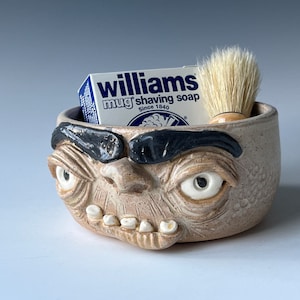 Puede incluir: Un tazón de afeitar de cerámica con forma de cara con una expresión de enfado. El tazón tiene cejas negras, ojos blancos y una sonrisa de dientes. Contiene una pastilla de jabón de afeitar Williams mug y una brocha de afeitar.