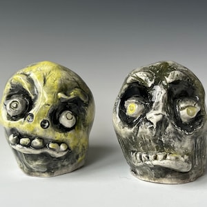 Peut inclure: Deux figurines de crâne en céramique aux traits exagérés. Un crâne est vert avec des détails noirs, l'autre est noir avec des détails verts. Les deux ont des yeux jaunes.