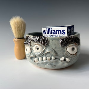Puede incluir: Un tazón de afeitar de cerámica con una cara de dibujos animados, que presenta dos ojos grandes, una boca ancha con dientes y un esmalte gris texturizado. Una barra de jabón de afeitar Williams mug está dentro del tazón, y un cepillo de afeitar está al lado.