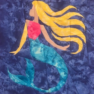 May include: A colorful paper craft of a mermaid with a blue tail, pink bodice, and long yellow hair. The mermaid is set against a blue background with a mottled pattern.