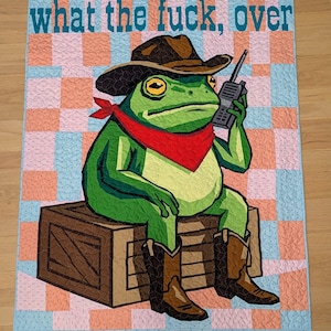 May include: A rectangular quilt featuring a green frog wearing a brown cowboy hat, a red bandana, and brown cowboy boots. The frog sits on a brown wooden crate while holding a black walkie-talkie. The text "what the fuck, over" is at the top.