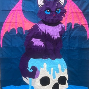 May include: A black cat with pink wings sits on a white skull with blue dripping paint. The cat has blue eyes and a purple and white fur pattern. The background is a dark blue.