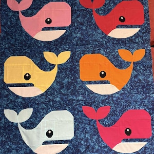 May include: A colorful quilt featuring six stylized whales in shades of pink, red, yellow, orange, and light blue. Each whale has a white belly and a black eye, set against a blue patterned background.