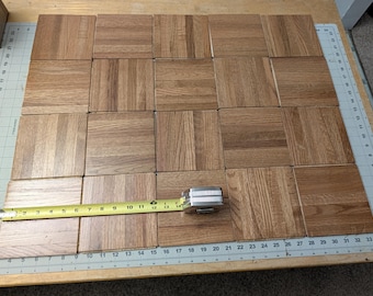 Puede incluir: Una cuadrícula de veinte baldosas cuadradas de madera de roble marrón claro dispuestas en un patrón de cuatro por cinco. Una cinta métrica metálica amarilla se extiende por la fila inferior, mostrando que las baldosas miden 35,5 cm de ancho.