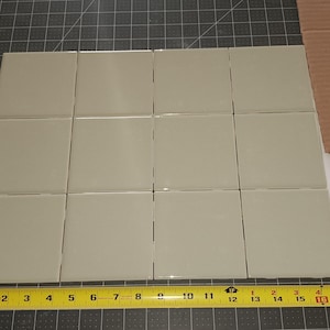 May include: Twelve square, light gray ceramic tiles arranged in a grid. A yellow measuring tape is placed along the bottom, showing measurements in inches. A piece of cardboard and white paper are in the background.