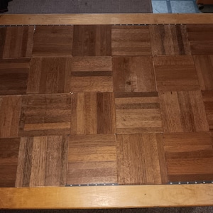 May include: A rectangular wooden parquet panel composed of twenty square tiles arranged in a four-by-five grid. The wood features a medium brown tone with visible grain patterns. A black measuring tape with white markings runs along the left edge.