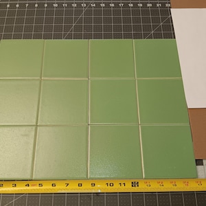 May include: A grid of twelve square sage green tiles arranged in a three-by-four pattern. A yellow measuring tape is positioned along the bottom edge, showing the total width of the four tiles is approximately 18 inches across the bottom.