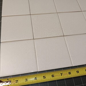 Puede incluir: Azulejos cuadrados blanquecinos dispuestos en forma de cuadr&iacute;cula. Una cinta m&eacute;trica amarilla muestra que los azulejos miden aproximadamente 10 cm de ancho. Los azulejos tienen una textura moteada y est&aacute;n sobre una esterilla de corte gris.