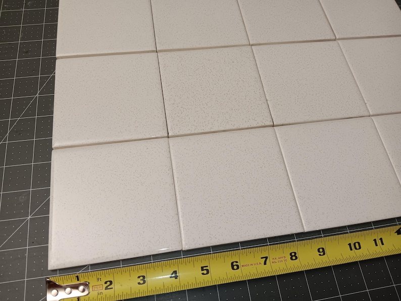 Puede incluir: Azulejos cuadrados blanquecinos dispuestos en forma de cuadr&iacute;cula. Una cinta m&eacute;trica amarilla muestra que los azulejos miden aproximadamente 10 cm de ancho. Los azulejos tienen una textura moteada y est&aacute;n sobre una esterilla de corte gris.