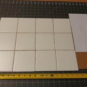 May include: A collection of sixteen white square tiles arranged in a grid, with a yellow measuring tape showing inches. A piece of white paper and a brown piece of cardboard are also present.