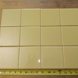 May include: A collection of twelve square, pale yellow tiles arranged on a wooden surface. A yellow measuring tape is placed below the tiles, showing measurements up to 17 inches. The tiles are likely for a home improvement project.