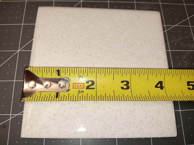 Puede incluir: Una baldosa cuadrada blanca con un patr&oacute;n moteado se mide con una cinta m&eacute;trica amarilla. La baldosa mide aproximadamente 10 cm de ancho. La cinta m&eacute;trica est&aacute; marcada en pulgadas y pies.