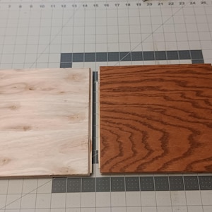 May include: Two square wooden panels. One is light-colored with a natural wood grain, the other is stained a rich brown with a prominent wood grain pattern. Both are on a gray cutting mat.