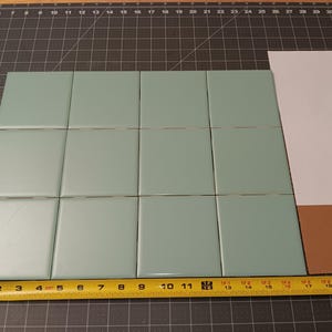 May include: A collection of twelve square, light teal tiles arranged in a grid. A yellow tape measure shows the tiles are approximately 10 inches wide. A white and brown paper is to the right of the tiles.
