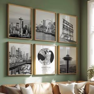 May include: Six framed black and white Seattle city prints hang on a green wall above a brown leather sectional sofa. The prints depict the Space Needle, a Ferris wheel, and a map of Seattle. The room is decorated with neutral-toned pillows and a white coffee table.