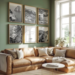 Puede incluir: Seis impresiones enmarcadas en blanco y negro de paisajes cuelgan de una pared verde sobre un sofá seccional de cuero marrón. Las impresiones representan escenas costeras y un mapa de Capri. Una mesa de centro blanca está frente al sofá.