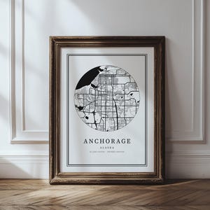 May include: A framed print showing a black and white circular map of Anchorage, Alaska. The city's name is printed below the map. The frame is a dark brown colour. The print includes latitude and longitude.