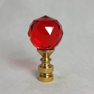 May include: A red crystal finial with a faceted design, mounted on a brass base. The finial is a decorative element used on lampshades, curtains, and other home decor items.