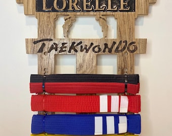 Op de afbeelding: Houten Taekwondo-gordelrek met de naam "Lorelle" gegraveerd op de bovenkant. Het rek heeft vijf vakken voor gordels, die zwart, rood, blauw, geel en wit zijn.