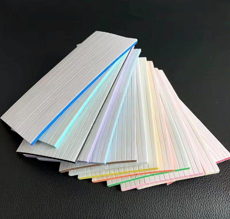 May include: A fan of colorful, textured, rectangular strips with various colored edges. The strips are arranged in a fanned-out pattern, showcasing a range of pastel and bright colors, including blue, green, pink, and yellow.