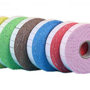 May include: A collection of colorful foam tape rolls. The rolls are in shades of yellow, green, brown, blue, red, and pink. Each roll has a white backing with small black dots. The tape is likely used for crafts or packaging.