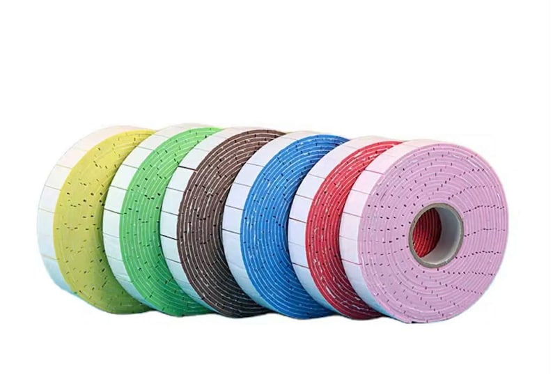 May include: A row of colorful rolls of double-sided adhesive tape. The rolls are in shades of yellow, green, brown, blue, red, and pink. Each roll has a white backing with small square adhesive sections.